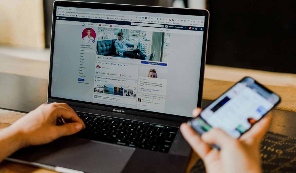 Hands Using Laptop with Screen Showing Facebook Page and Holding Cell Phone