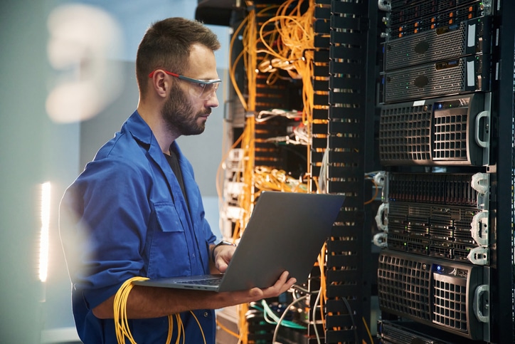 Silver colored laptop in hands. Young man is working with internet equipment and wires in server room.